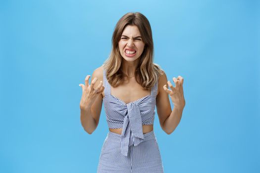 Indoor shot of irritated moody woman having mental breakdown feeling annoyed and pressured squeezing hands in fists from anger wrinkling nose in hate standing hateful over blue background. Emotions concept