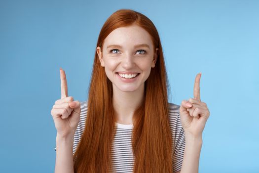 Amused excited happy smiling redhead girl look pointing up grinning gladly watching awesome open air perfomance standing joyfully showing cool thing indicating product, blue background.