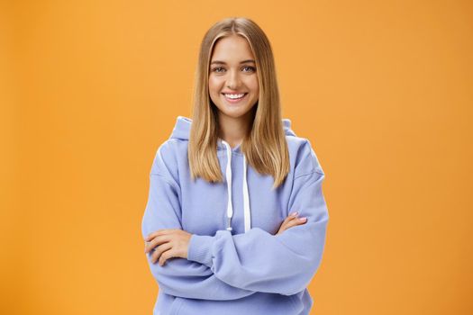 Young creative and ambitious female student staring own project at home working freelance having free time and working when want holding hand crossed on chest wearing cozy hoodie over orange wall. Lifestyle.