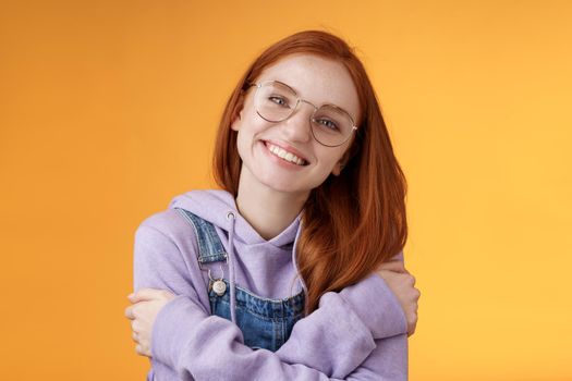 Tender romantic european redhead girlfriend tilting head cute lovely smile white teeth tilting head hugging herself wearing best friends hoodie adore new outfit grinning joyfully, orange background.