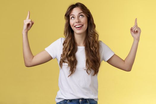 Happy admiring cute european curly-haired girl long haircut look pointing up impressed amused smiling broadly satisfactory delighted stand white t-shirt yellow background joyfully react good promo. Lifestyle.