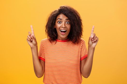 Waist-up shot of amazed happy enthusiastic cute african american woman with curly hairstyle in striped t-shirt raising index fingers pointing up gazing mesmerized and delighted at camera over orange wall. Lifestyle.