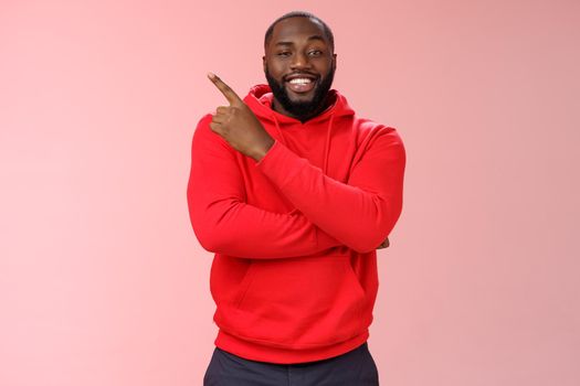 Cheeky outgoing smiling attractive african-american male model in red hoodie grinning energized pointing upper left corner indicating awesome product recommend promo, standing pink background.