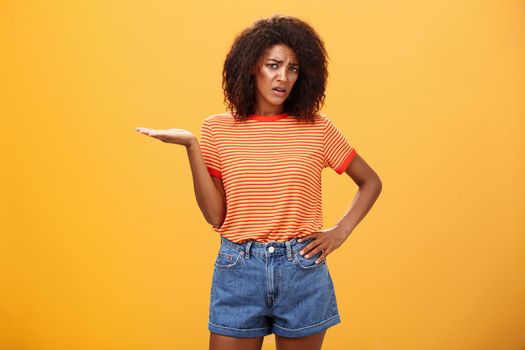 So what. Portrait of moody unimpressed stylish dark-skinned girlfriend with curly hairstyle holding hand on waist frowning and raising palm in your point gesture being confused and questioned over orange wall.