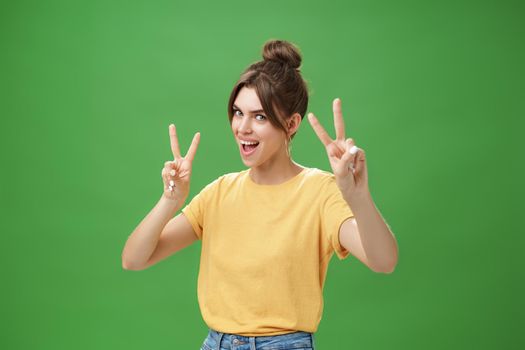 Indoor shot of enthusiastic excited and happy daring girl with combed hair tattoo and cute diasdema showing peace signs bending backwards standing in cool energized pose over green background. Lifestyle.