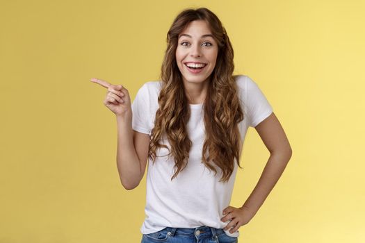 Lively surprised happy enthusiastic good-looking woman long curly hairstyle white t-shirt laughing impressed astonished pointing left index finger discuss interesting exhibition yellow background.