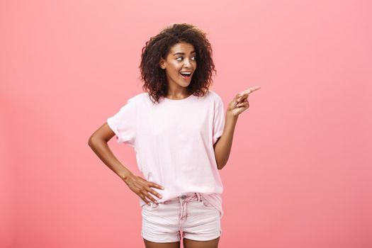 Hey wanna go there. Portrait of intrigued pleased good-looking stylish african woman on vacation wearing summer clothes pointing and gazing right with curious look over pink background. Advertisement concept