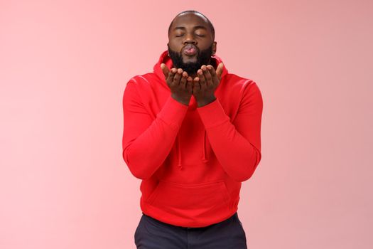 Charming cute african american bearded guy in red hoodie close eyes folding lips hold palms near mouth sending passionate air kiss blowing mwah camera, standing lovely pink background.