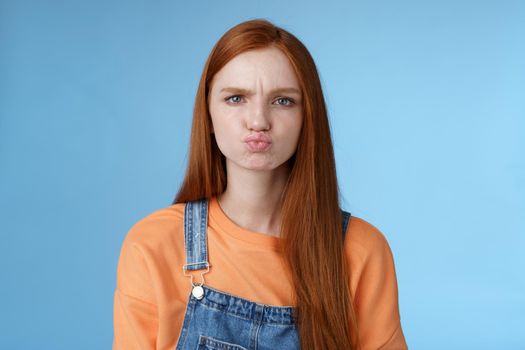 Moody displeased clingy girlfriend redhead blue eyes pouting sulking upset offended frowning making grimace showing attitude standing disappointed unsatisfied blue background, complaining.