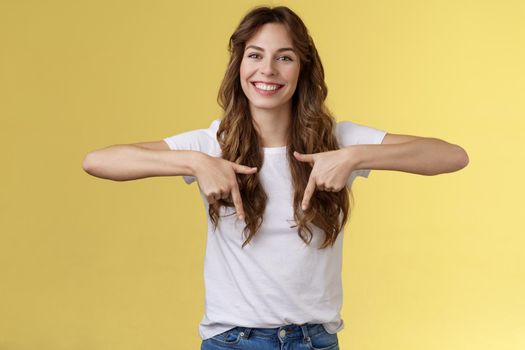 Lively helpful good-looking caucasian girl curly long hair showing you excellent place find hairdresser pointing down index fingers smiling toothy happy delighted grin stand yellow background. Lifestyle.