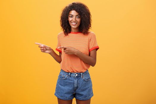 Portrait of charming friendly-looking african-american female coworker in casual outfit pointing left and smiling broadly showing perfect copy space for customers against orange background. Lifestyle.