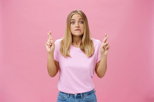 Woman hopefully looking at chart praying with crossed fingers to pass exam biting lip popping eyes, standing intense and worried against pink background having hope to win or receive scholarship. Body language concept