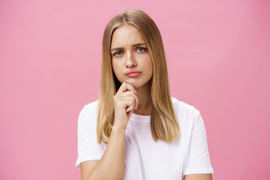 Girl facing problem thinking being hesitant while making decision holding hand on chin frowning pursing lips standing thoughtful over pink background in white casual t-shirt. Body language concept