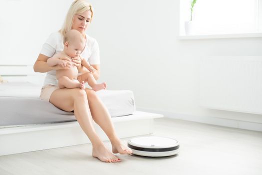 Young mother sitting on sofa with toddler son and watching robot vacuum cleaner doing housework