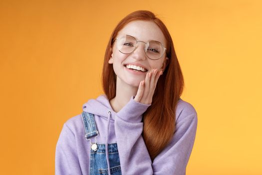 Charismatic happy tender redhead smiling european girl 20s glasses hoodie denim overalls having fun enjoying summer holidays chilly evening laughing joyfully touch pure clear skin, orange background.