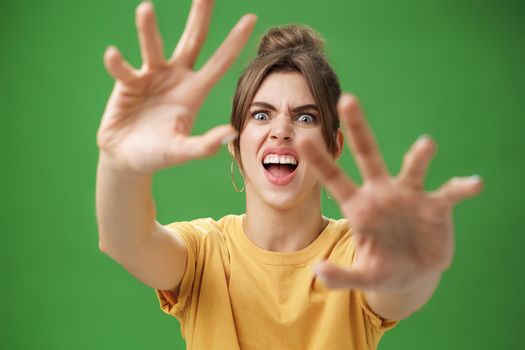 Funny emotive woman making fun face pulling hands forward to attack of grab something frowning opening mouth wanting cover face from camera, grimacing over green background. Body language and emotions concept