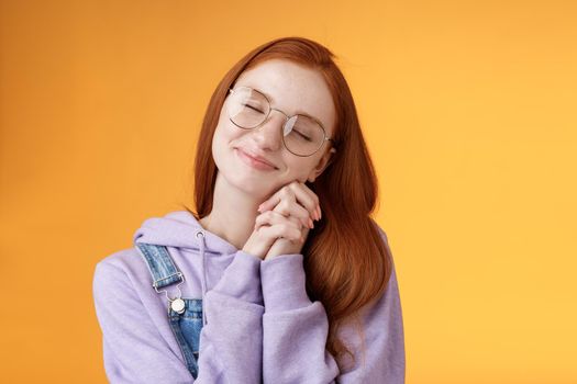 Sweet tender happy romantic redhead girlfriend close eyes smiling dreamy imaging lovely scene daydreaming press palms cheeks silly recalling nice memories, standing orange background relaxed.