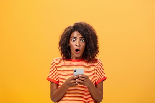 Portrait of shocked and concerned stunned dark-skinned woman with afro hairstyle popping eyes from concern and panic opening mouth holding smartphone revealing terrible information.
