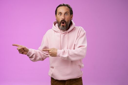 Astonished fascinated curious mature bearded man with grey hair in pink hoodie folding lips amazed pointing left index fingers widen eyes camera impressed thrilled wanna go check out cool promo. Lifestyle.