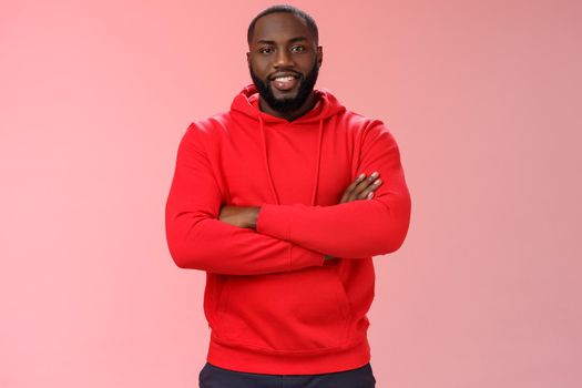 Attractive bearded african-american young guy wearing red hoodie cross arms chest smiling friendly showing friends new place grinning delighted buying own flat, standing proud pink background.