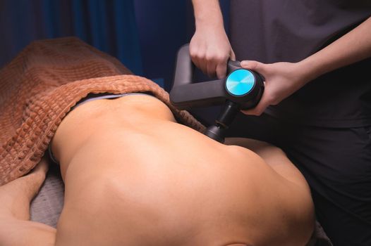 Close-up of a professional male masseur stimulates the back muscles of a male patient in a dark spa room for massage. Percussion mechanical effect on overstrained muscles.