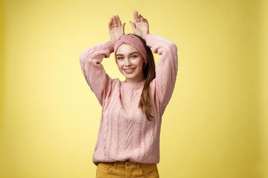 Nice charming playful young european female having fun, playing mimicking bunny showing rabbit ears with hands behind head, smiling cute, tender, happy greeting boyfriend welcome back ove yellow wall.