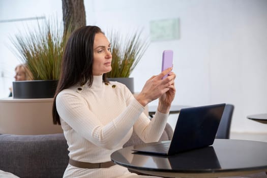 beautiful mid adult woman typing smartphone sitting by laptop in public place