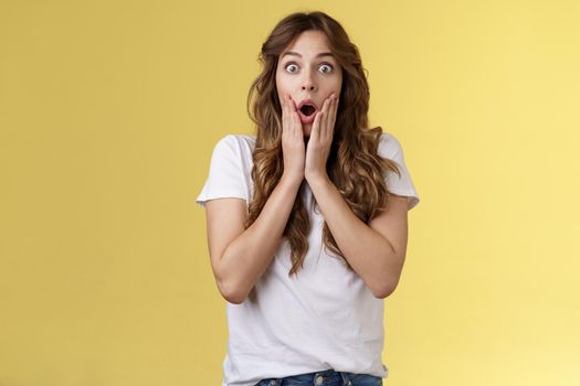 Shocked speechless impressed curly-haired silly girl drop jaw gasping astonished grab cheeks stare camera interested surprised checking out incredible unexpected scene yellow background.