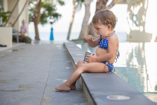 portrait of little girl eating ice cream yogurt with spoon sitting by outdoor pool