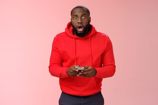 Amused overhwelmed black bearded guy in red hoodie drop jaw gasping widen eyes staring camera impressed holding smartphone using amazing cool game astonished new phone features, standing pink wall.