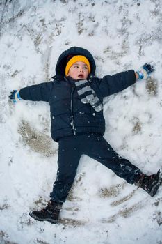 The boy in a blue jacket and blue pants lies in the snow and makes an angel. His yellow hat comes to the fore. The play and joy of a child in the snow.