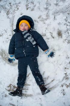 The boy in a blue jacket and blue pants lies in the snow and makes an angel. His yellow hat comes to the fore. The play and joy of a child in the snow.