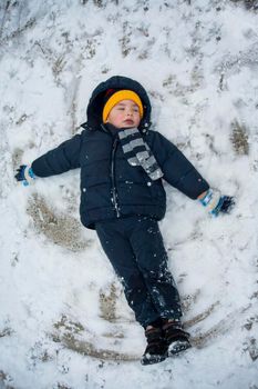 The boy in a blue jacket and blue pants lies in the snow and makes an angel. His yellow hat comes to the fore. The play and joy of a child in the snow.