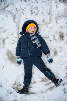 The boy in a blue jacket and blue pants lies in the snow and makes an angel. His yellow hat comes to the fore. The play and joy of a child in the snow.