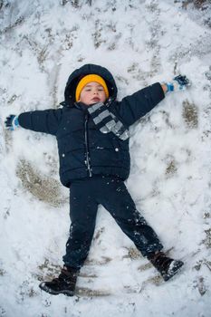 The boy in a blue jacket and blue pants lies in the snow and makes an angel. His yellow hat comes to the fore. The play and joy of a child in the snow.