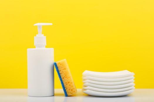 Colorful dishwashing concept, creative composition with liquid detergent in white plastic bottle with dispenser, yellow cleaning sponge and pile of clean plates against yellow background