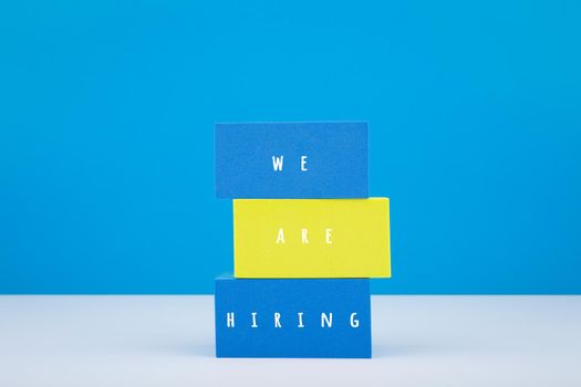 Trendy job opening concept. We are hiring written on colorful blue and yellow blocks on white table against blue background with copy space