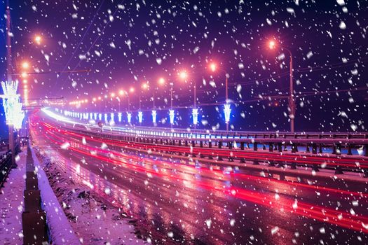 Traces of headlights from cars moving at winter night on the bridge, illuminated by lanterns in a snowfall. Lights reflecting in the wet asphalt.