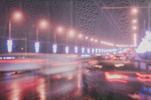 Blurry lights of moving cars and lanterns reflecting on the wet asphalt in the night city behind rain-covered window. Rainy bad autumn weather concept.