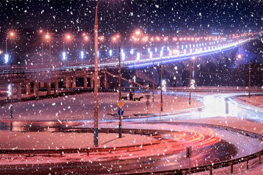 Traces of headlights from cars moving at winter night on the bridge, illuminated by lanterns in a snowfall. Lights reflecting in the wet asphalt.