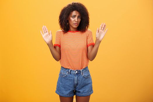 Silly insecure and sad dark-skinned female model in trendy striped t-shirt and shorts raising arms in surrender frowning being uninvolved and unaware standing clueless over orange background. Lifestyle.
