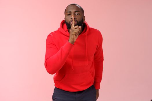 Shhh it taboo. Portrait sensual funny cute african-american bearded man in red hoodie bending camera show shush gesture index finger on lips telling secret asking keep quiet not tell anyone mystery.