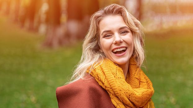 Joyful young woman in autumn coat and yellow knitted scarf standing joyful smiling in the fall yellow garden or park. Beautiful smiling young woman in autumn leaves.
