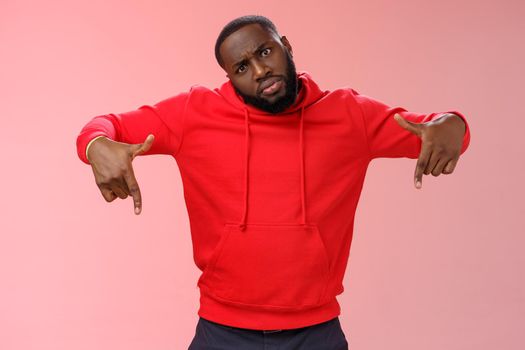 Cheeky stylish good-looking black bearded guy look cool tilting head bossy confident frowning seriously pointing down showing awesome place hang out homies, standing pink background.