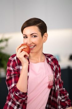 Smelling fresh apple holding it in a hand, with a sexy eye contact on camera charming housewife wearing plaid shirt with a short hairstyle while cooking apple pie standing at the kitchen.