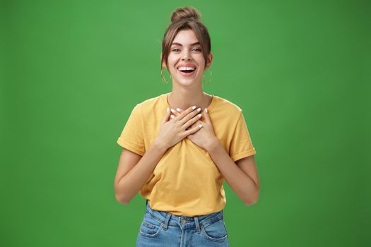 Delighted charming woman with gapped teeth in cozy outfit holding palms on chest in grateful or thankful pose smiling broadly receiving warm congratulations standing satisfied, happy over green wall. Body language and emotions concept