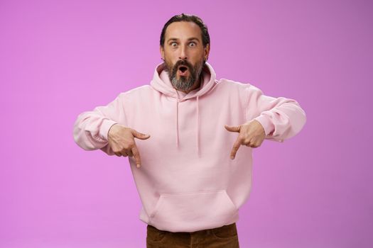 Impressed fascinated speechless dad bearded grey hair say wow widen eyes surprised folding lips amused curiously pointing down index fingers see super amazing advertisement, posing purple background.