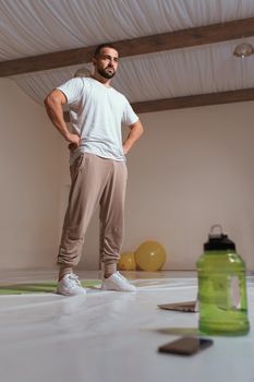 Strong athletic handsome man in white t-shirt and sweatpants taking a break standing at hall in his apartment. Pensive sportsman with thoughtful face ponders workout program he found online.