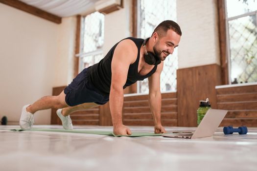 Young sport man doing up push ups exercise in empty gym or home watching online sports videos. Muscular sportsman doing exercises alone while self distance or lockdown in gym.
