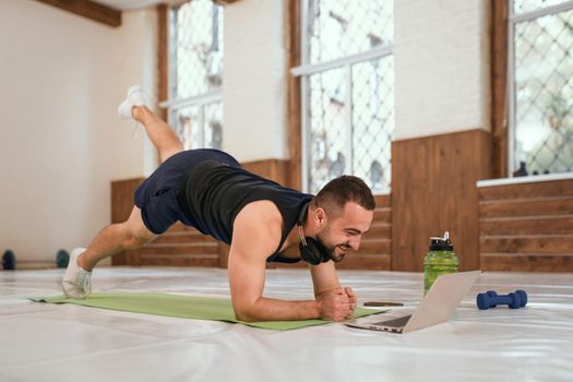 Handsome young sport man doing one leg up push ups exercise in empty gym or home watching online sports videos all in black clothes. Muscular sportsman doing exercises alone in gym.